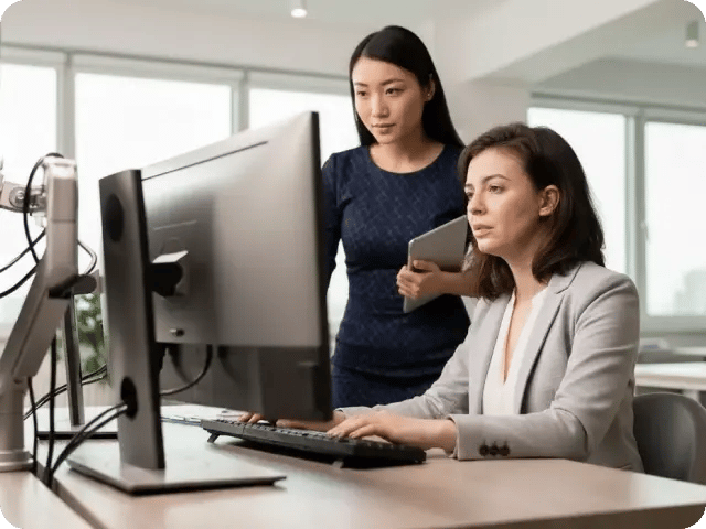 A professional woman standing and collaborating with a seated colleague working at a computer.