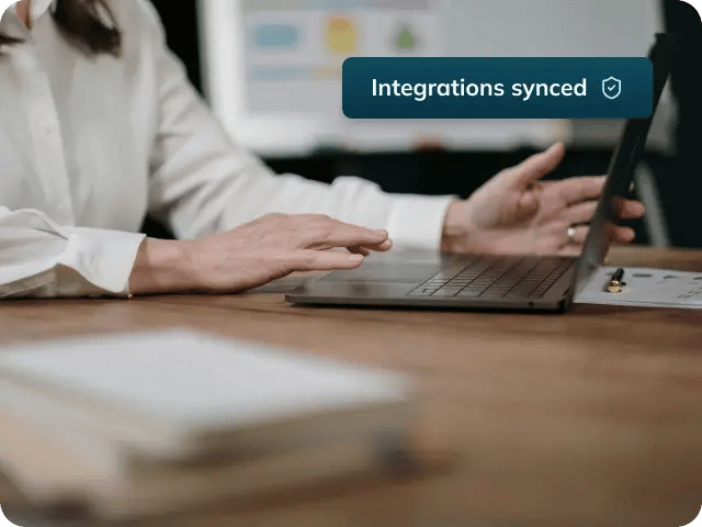 A woman working on a laptop with a 'Systems synced' UI snippet featuring a shield and check icon in the top-right corner.