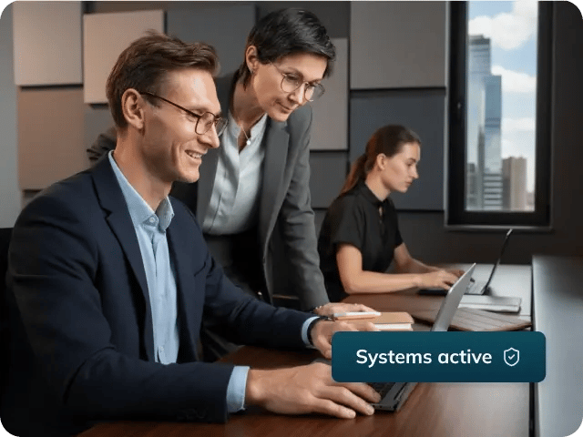 Three people working in an office setting with a 'Systems active' UI snippet and shield icon in the bottom right.