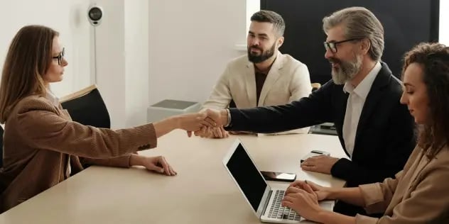 A group of professionals in a boardroom meeting, featuring two people shaking hands to signify an agreement.