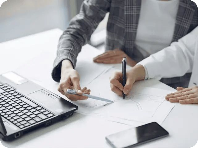 A close-up of two people's hands at a work table, using pens to point at and annotate a piece of paper with a line graph.
