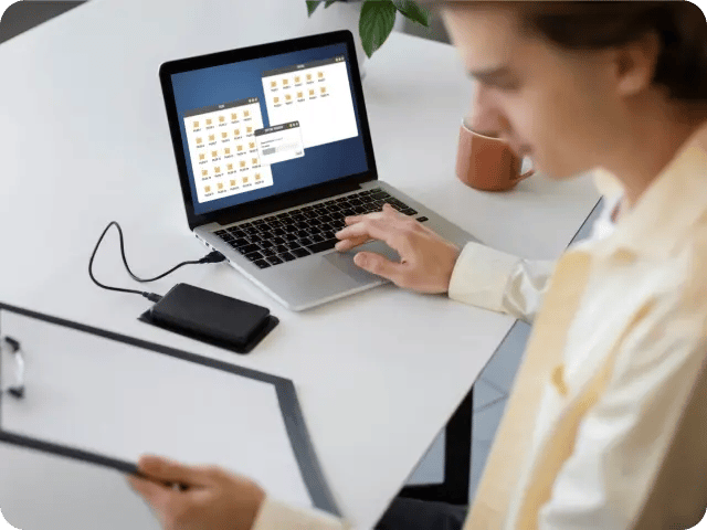 A woman using a laptop with an external drive connected, while reviewing a document on a clipboard.